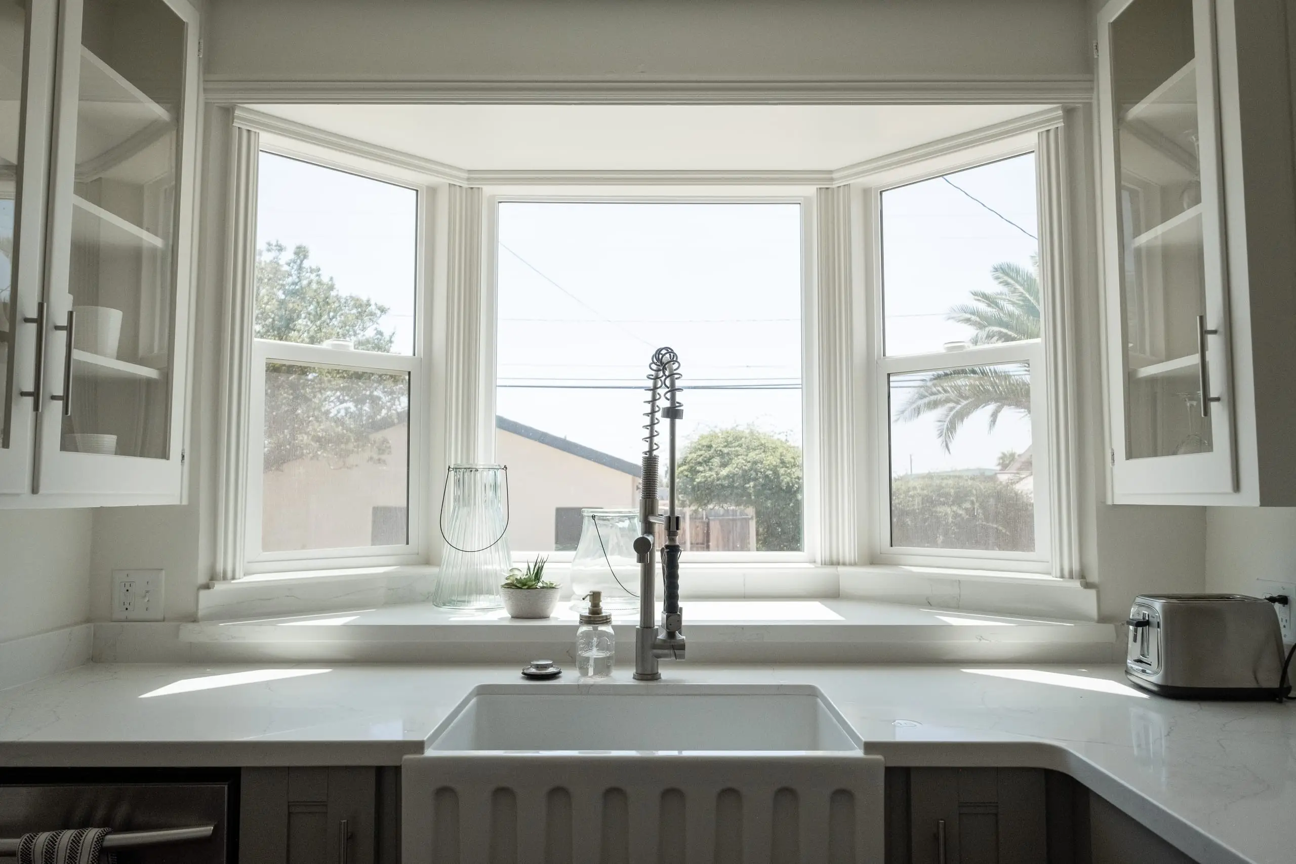 A bright kitchen with a white farmhouse sink and a tall chrome mixer tap under a bay window